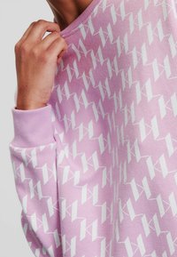 Ljust rosa sweatshirt med vitt geometriskt mönster, med ribbade muddar och halsringning, visas hållas i en hand.