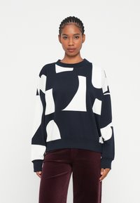 Sudadera azul marino con grandes formas abstractas blancas, cuello redondo y puños acanalados. Combinada con pantalones de pana burdeos.