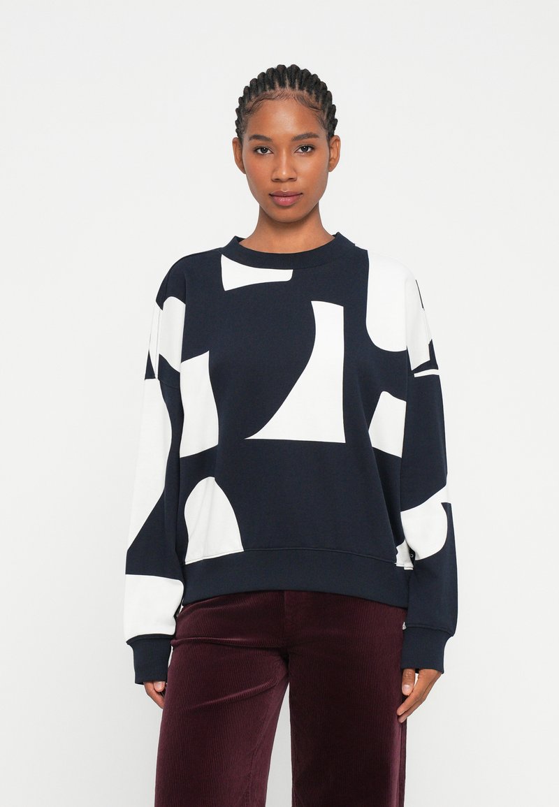 Sudadera azul marino con grandes formas abstractas blancas, cuello redondo y puños acanalados. Combinada con pantalones de pana burdeos.