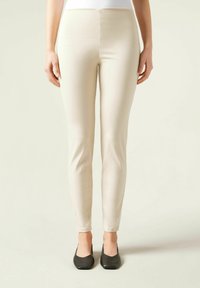 Leggings beige ajustés en tissu extensible, présentant une texture lisse et une taille haute, assortis à des chaussures plates noires.
