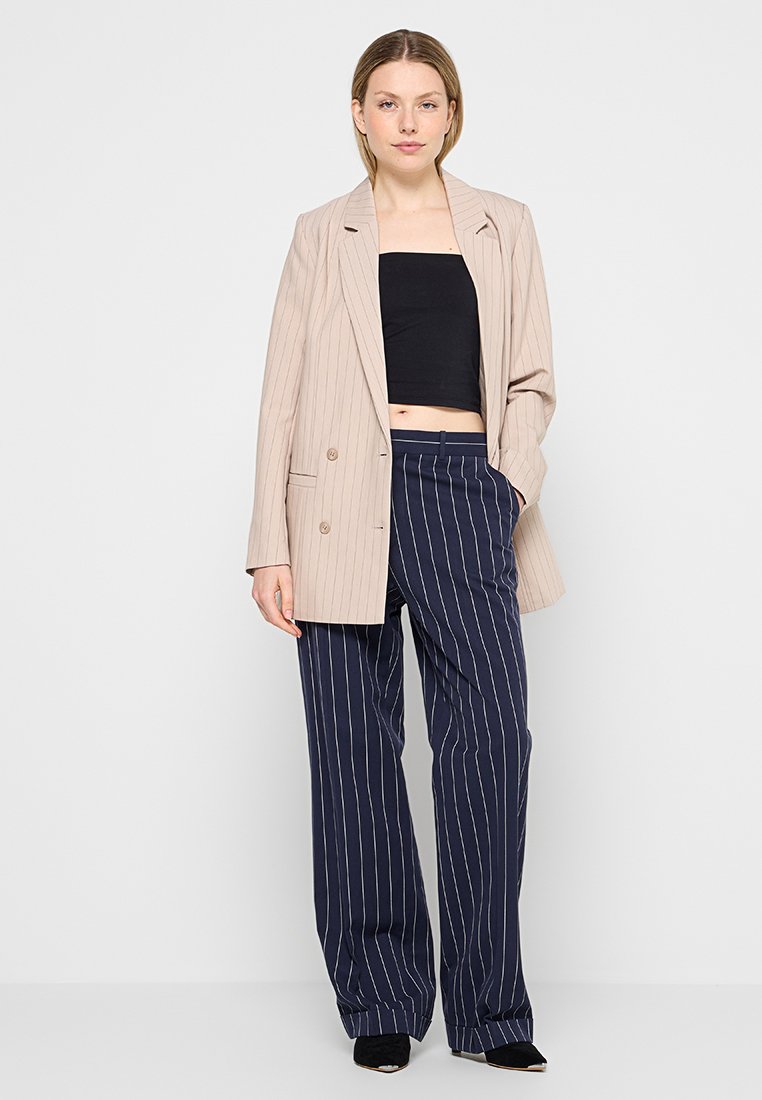 Femme portant un blazer beige à rayures, un crop top noir sans manches, un pantalon large bleu marine à rayures, et des chaussures noires à talons pointus, se tenant avec une main dans la poche.
