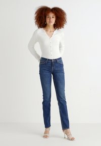 Top bianco a maniche lunghe con scollo a V e bottoni abbinato a jeans in denim blu a vita alta e tacchi bianchi con cinturini.