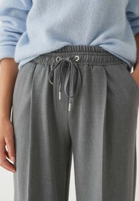 Grey tailored trousers with a pleated design, elastic waistband, and drawstring closure featuring metal tips. Soft fabric texture.