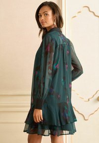 Rochia fluidă de culoare teal, cu mâneci transparente, guler negru și hem învolburat în straturi. Prezintă un model floral în tonuri de purpuriu și verde.