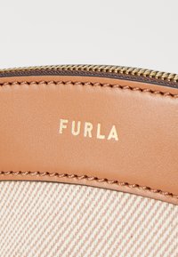 Detail van een bruine leren tas met zichtbare stiksels, een gouden rits en een beige gestreepte stof waarin de naam "FURLA" in het goud is geperst.