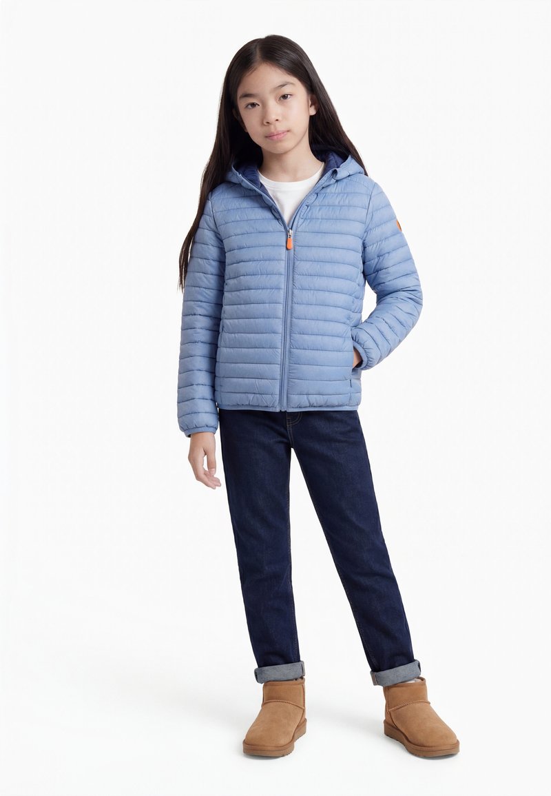 Νεαρή κοπέλα που στέκεται φορώντας μια ανοιχτό μπλε βαμβακερή μπουφάν, σκούρα τζιν γυρισμένα στην κορυφή και τα καφέ μποτάκια στον αστράγαλο σε λευκό φόντο.