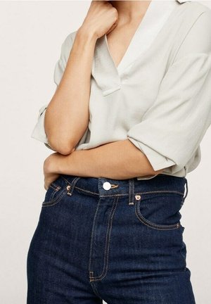 Personne portant une chemise beige clair à manches longues avec les manches retroussées et un jean taille haute bleu foncé, bras croisés et main près du menton.