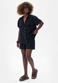 Zwarte linnen shirt met korte mouwen en twee voorzakken, gecombineerd met bijpassende shorts. Model draagt donkere zonnebrillen en bruine platform sandaal.