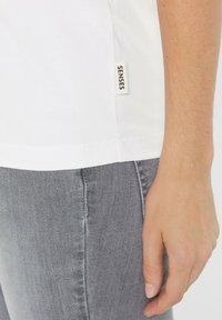 Weißes T-Shirt mit kurzen Ärmeln und einem Seitenetikett mit der Aufschrift "SENSES", kombiniert mit grauen Jeans. Glatte Textur und einfache Designdetails.