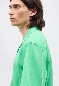 Νέος με καστανά μαλλιά μέχρι τους ώμους, φοράει χαλαρό πράσινο πουκάμισο, κοιτά προς τα αριστερά μπροστά σε απλό φωτεινό φόντο.