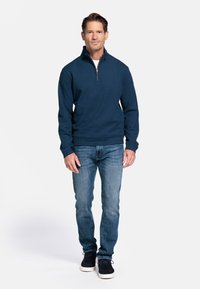 Marine blazer met ritssluiting tot de helft, gemaakt van gestructureerde stof, gecombineerd met lichtblauwe spijkerbroek en donkere sneakers. Model staat tegen een effen achtergrond.