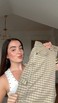 Fiatal nő, aki hosszú sötét hajat visel és fülbevalót, egy bézs és szürke kockás szoknyát tart a kezében egy szobában, ahol egy arany csillár lóg.