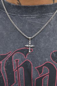 Collana in argento con pendente a croce decorato con dettagli a forma di teschio, appoggiata su una camicia grigia scura con texture e stampa grafica rossa.