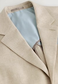 Blazer beige con tejido texturizado, forro azul claro, solapas con muesca y detalles de costura sutiles. Incluye un lazo colgante en el interior.