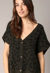 Cardigan tricoté présentant un motif léopard vert foncé, un col en V, des manches courtes et cinq grands boutons sur le devant. Tissu doux et texturé.