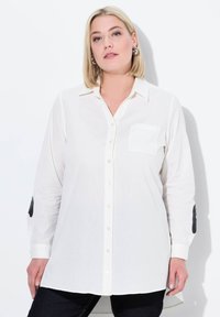 Camicia bianca a maniche lunghe con bottoni, dotata di una tasca sul petto e toppe nere sui gomiti. Tessuto morbido con una vestibilità rilassata e orlo curvo.