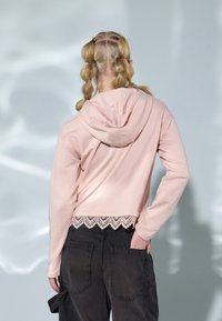 Skrácená růžová mikina s kapucí a krajkovým lemem dole, spárovaná s tmavě texturovanými cargo kalhotami s velkými kapsami.