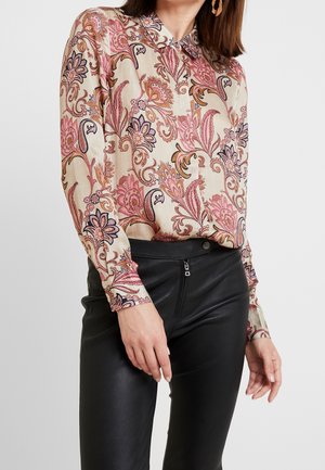 Vrouw met een beige blouse met lange mouwen en roze bloemenpatroon, ingestopt in een zwarte leren broek met hoge taille en een ritssluiting.