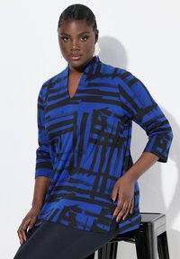 Blaue Tunika mit schwarzen abstrakten Mustern, V-Ausschnitt, dreiviertel Ärmel, seitliche Schlitze und leichtem Stoff. Getragen mit dunklen Leggings.