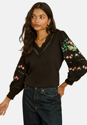 Schwarzer Pullover mit V-Ausschnitt und Spitzenbesatz. Lange Ärmel mit bunter Blumenstickerei und Lochdetails. Kombiniert mit dunklen Jeans.