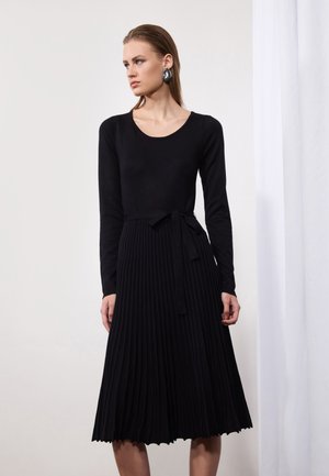 Black dress featuring a fitted, long-sleeve top and a pleated, A-line skirt. Complements include a tied waist and textured fabric.