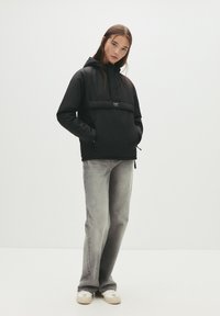 Sort letvægtsjakke med hætte og frontlomme, parret med falmede grå jeans og hvide sneakers. Enkel, funktionel design.