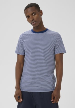 Ung mand med kort hår iført en blå og hvidstribet kortærmet skjorte og mørke shorts mod en ensfarvet baggrund.