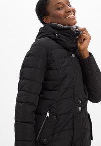 Zwarte pufferjas met een hoge kraag, zachte fleece-details, drukknopen aan de voorkant en een ritszak aan de linkerkant.