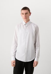 Camisa blanca de manga larga, hecha de tejido suave, con un cuello clásico, cierre frontal con botones y dobladillo redondeado. Se lleva con pantalones oscuros.