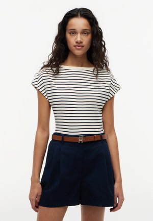 Donna con lunghi capelli ricci scuri che indossa una maglietta a maniche corte a righe bianche e blu navy, pantaloncini blu navy e una cintura marrone con fibbia argentata.