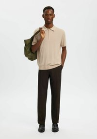 Homme portant un polo beige à manches courtes et un pantalon foncé, tenant un sac vert olive sur l'épaule, debout devant un fond blanc.