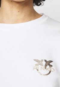 Valkoinen collegepaita, jossa on pyöreä pääntie ja kultainen brodeeraus kahdesta linnusta sanan "PINKO" alla vasemmassa rintapielessä.