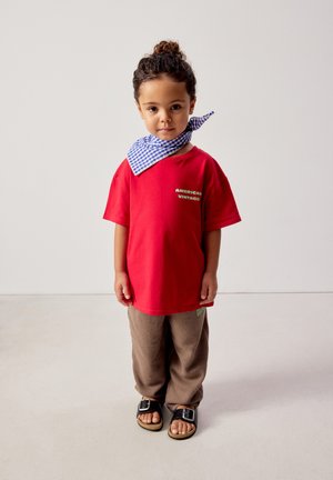 Kind met gekruld haar in een staart, draagend een rood shirt van "American Vintage", bruine broek, zwarte sandalen en een blauw geruiten nekdoek, staand op een witte vloer.