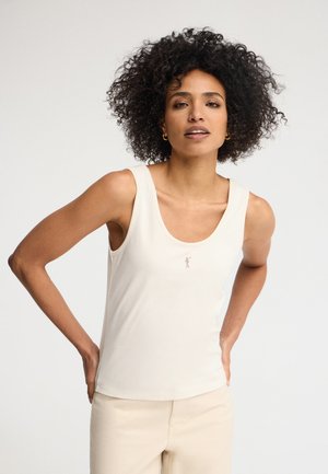 Kobieta o kręconych włosach, ubrana w białą, bezrękawnikową bluzkę i jasne spodnie, stojąca z rękami na biodrach na tle jednolitego tła.