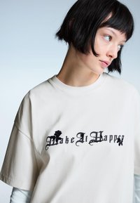 Crèmekleurig oversized t-shirt met zwarte tekst "Make It Happy" met bloemaccenten. Effen stof, korte mouwen, ronde hals.