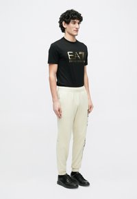 Juoda marškinėliai su auksiniu logotipu, kremi spalvos sportinės kelnės su šoniniu logotipo spaudiniu ir juodi sportbačiai. Modelis stovi šonu prieš paprastą foną.