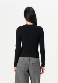 Eine Frau mit langen dunklen Haaren trägt ein enges schwarzes Langarmshirt und lockere graue Hosen und steht mit dem Rücken zu einem schlichten weißen Hintergrund.