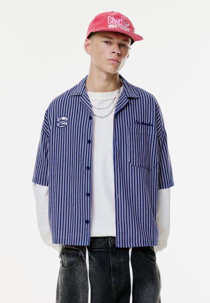 Gestreept shirt met korte mouwen in marineblauw en wit met een borstzak, laag over een wit shirt met lange mouwen, gecombineerd met donkere jeans en een roze pet.