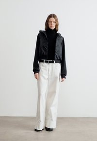 Γυναίκα με μαλλιά έως τους ώμους, φορώντας μαύρο ζιβάγκο, μαύρο αμάνικο γιλέκο, λευκά παντελόνια με φαρδιά μπατζάκια, μαύρη ζώνη και μαύρα παπούτσια.