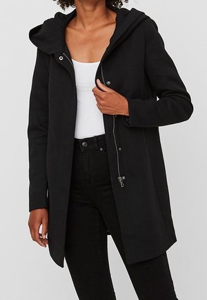 Kobieta w czarnym płaszczu z kapturem, zamkiem błyskawicznym i zatrzaskami, w połączeniu z białym topem i czarnymi spodniami na jednolitym tle.