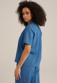 Zila denima krekls ar īsām piedurknēm, brīva silueta, ar taisnu apakšu un mīkstu tekstūru. Valkāts kopā ar atbilstošām zilajām biksēm.