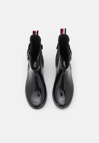 Bottines noires en cuir verni avec une finition lisse et brillante, dotées d'une doublure en tissu, d'une fermeture zippée et d'une languette de tirage rayée rouge et blanche.
