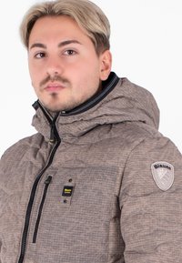Giubbotto imbottito marrone con cappuccio, dotato di tasca sul petto, zip nere e una toppa con logo sulla spalla. Materiale testurizzato.