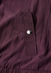 Diep bordeauxkleurige stof heeft een klein zakje met een zilveren drukknop, benadrukt door subtiele textuur en stikdetails.