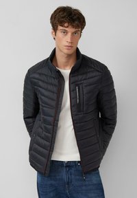 Gewatteerde zwarte jas met een opstaande kraag, horizontale stiksels, een borstzak met rits en een contrasterende rode ritssluiting.