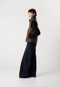 Kvinne med kort hår som har på seg en brun puffervest, svart kabelstrikkegenser og vide mørkeblå jeans, står sidelengs med hendene i lommene.