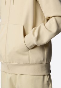 Haut de survêtement zippé beige en matériau doux, avec une poche kangourou, des poignets et un ourlet côtelés, une texture lisse et une coupe décontractée.