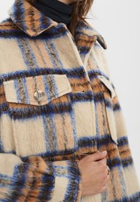 Nahaufnahme einer Person in einer beigen, blauen und braunen karierten Wolljacke mit geknöpften Brusttaschen, die die Jacke in der Nähe der Taille hält.