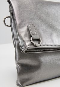 Metallinhohtoinen hopeanvärinen käsilaukku, jossa on sileä, teksturoitu pinta. Siinä on taitettavaa läppää ja sivulla pyöreä metallirengas.