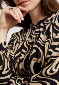 Svart och beige mönstrad blus med långa ärmar, rundad krage och knappstängning. Tygerna har en flödande textur med vågiga mönster.